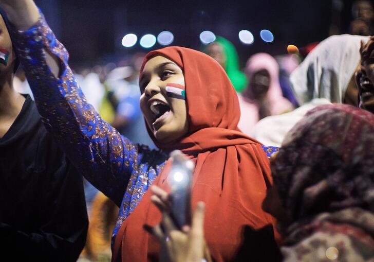 A woman in a red hijab and Sudanese flag on her face raises her hand in the air