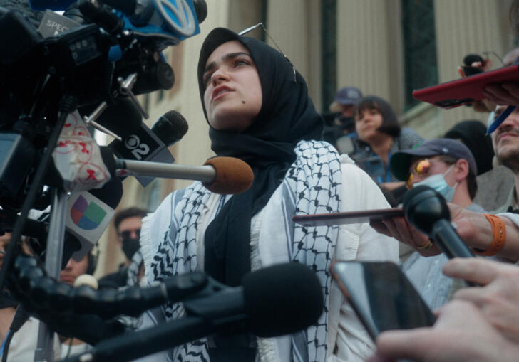 A woman wrapped in a kefiyah speaks into a camera and surrounded by journalists