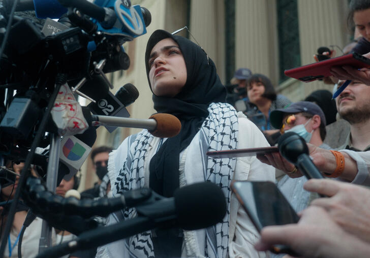 A woman wrapped in a kefiyah speaks into a camera and surrounded by journalists