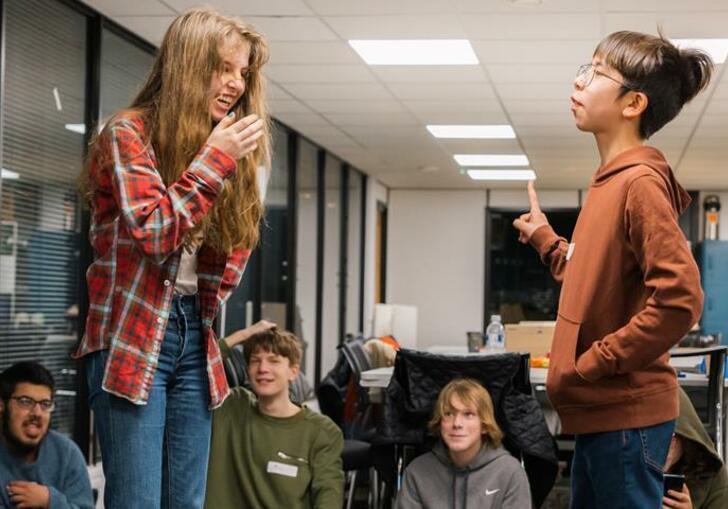 A group of children watch two others perform, one laughs as the other holds character.