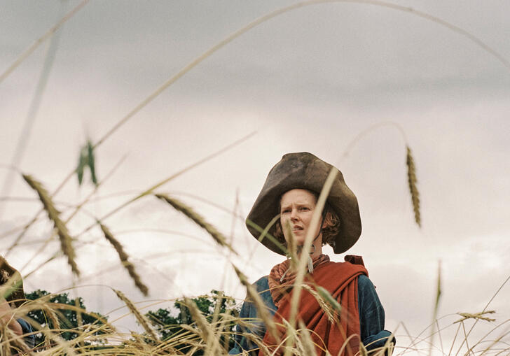 A person wearing a large leather hat and traditional Scottish tartan clothes stands obscured in a wheat field