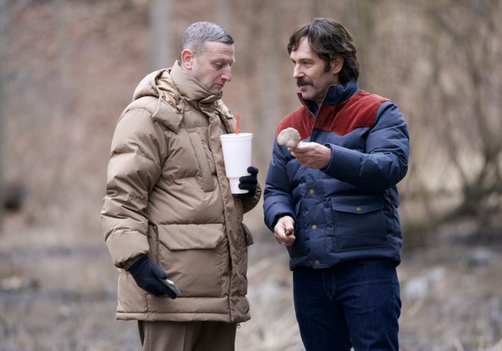 Two men in big winter coats share snacks on a walk