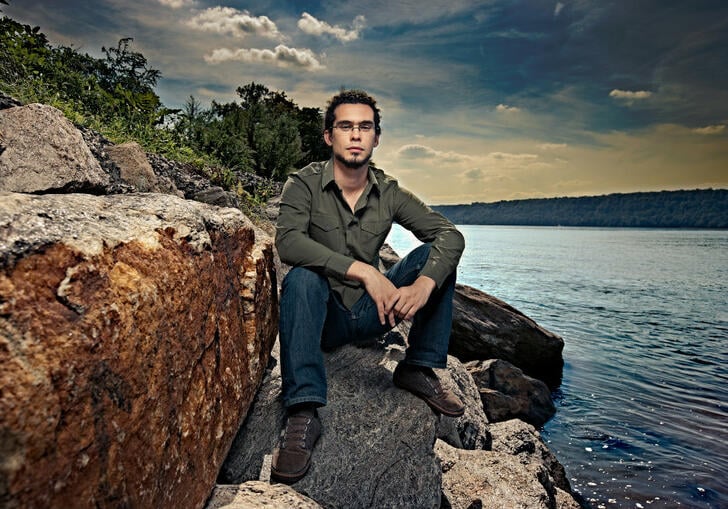 Jazz pianist and composer Fabian Almazan sits on large rocks by a serene lakeside, wearing a green shirt and jeans, with trees and a dramatic sky in the background.