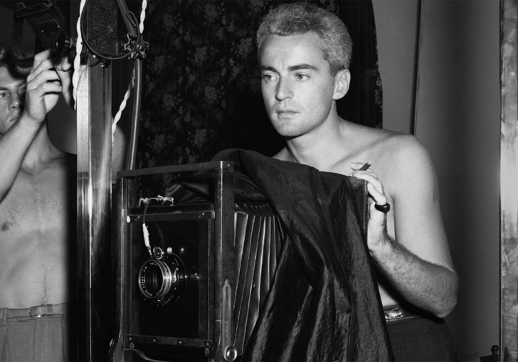 Two men stand behind an old fashioned camera, shirtless