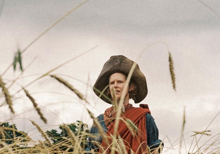 A woman wrapped in a red cloth and wearing a large leather hat stands in a field of wheat