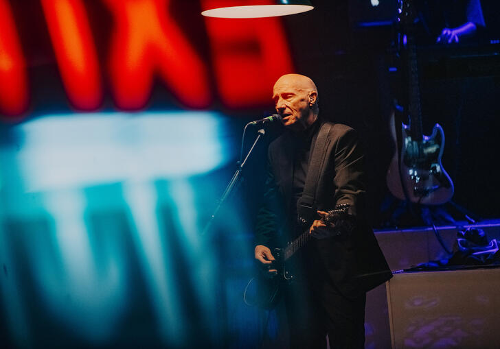 a photo of Midge Ure in a suit playing the guitar under the haze of a red and blue neon sign. 