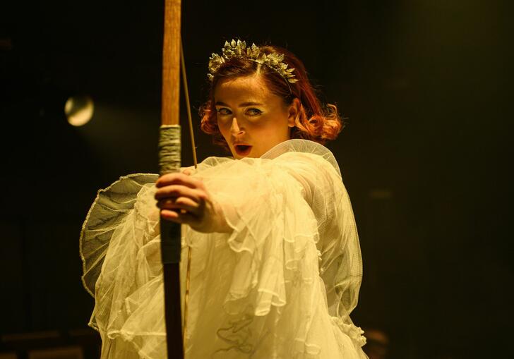 Opera singer dressed in white ruffled outfit and roman crown holding bow and arrow