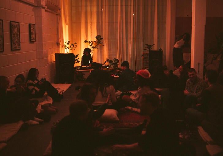 A group of people are gathered in a room with ambient lighting around a DJ