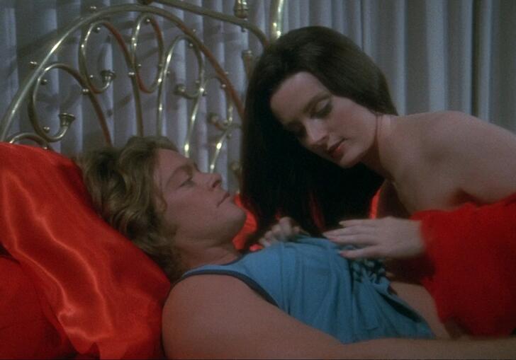 A woman lies in bed with a man, wrapped in red bed sheets. 