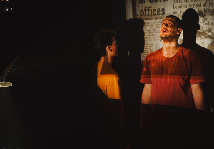 Two individuals are positioned in a dimly lit room. Image of newspaper text is projected behind them. Shadows are cast on the wall.
