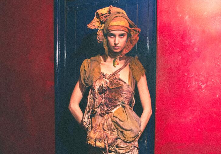 A model stands in front of a blue door and red wall, wearing earth toned distressed clothing