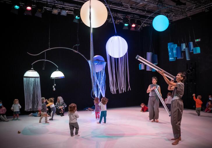 Toddlers running around with orbs of light hanging from the ceiling while two people overlook them