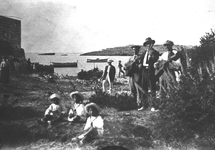 Black and white photo of Malta. There are three mail musicians performing and three children in the foreground. In the background is the ocean with some boats in it.