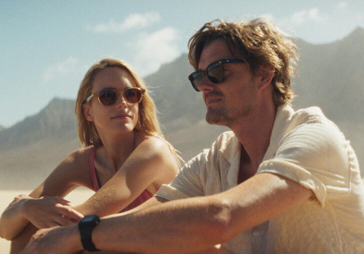 A man and a woman sit on the beach looking at each other with mountains in the background