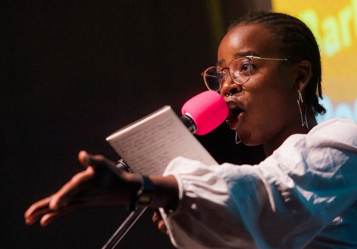 A young Black woman performing on stage speaking into a pink microphone, with her arm extended as she holds a book or paper.