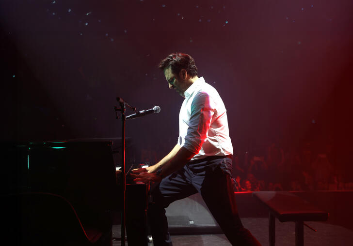 Image of Guy Manoukian on stage standing and paying the piano with a mic next to him