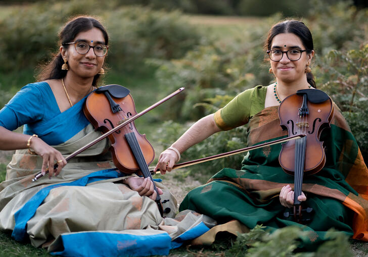 A photo of sibling duo Mathangi & Sambavi Uthayakumar sitting with violins on the grass.