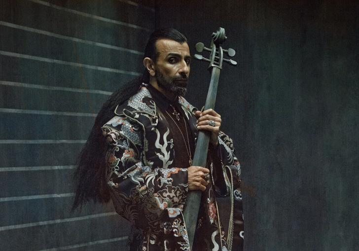 Ara Malikian with his hair tied back, in a long patterned robe holding a cello