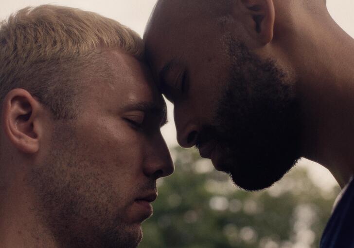 Two men stand together with their foreheads touching and eyes closed