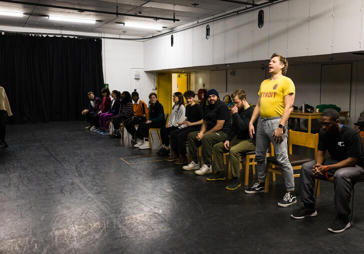 A group of workshop participants sit in a line of chairs. One of them is standing up and performing