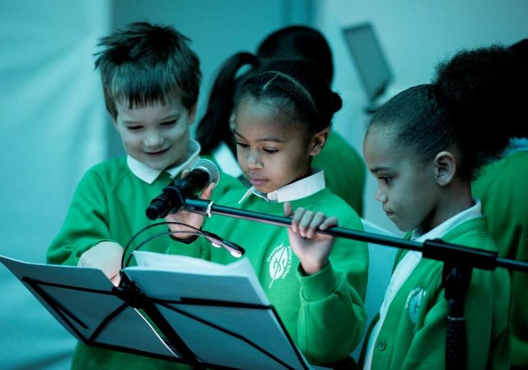 Photo of three young children by a microphone looking at sheet music