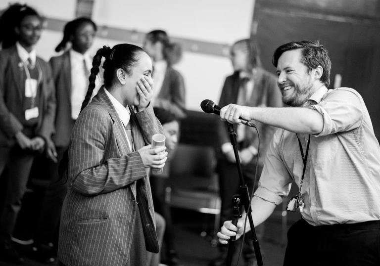Photo of girl in school uniform laughing as teacher holds microphone