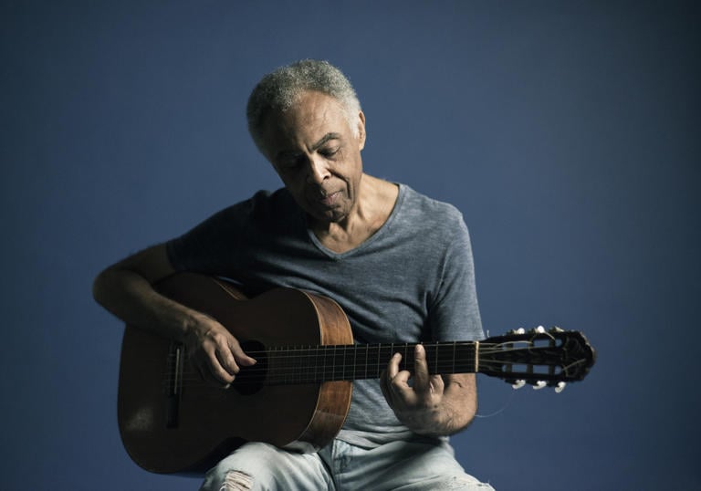 Gilberto Gil looking blue whilst playing his guitar in blue jeans and a blue t-shirt in front of a blue background