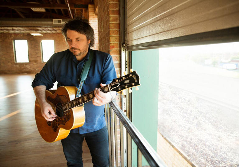 Jeff Tweedy playing guitar in the corner of a large room with exposed brick walls