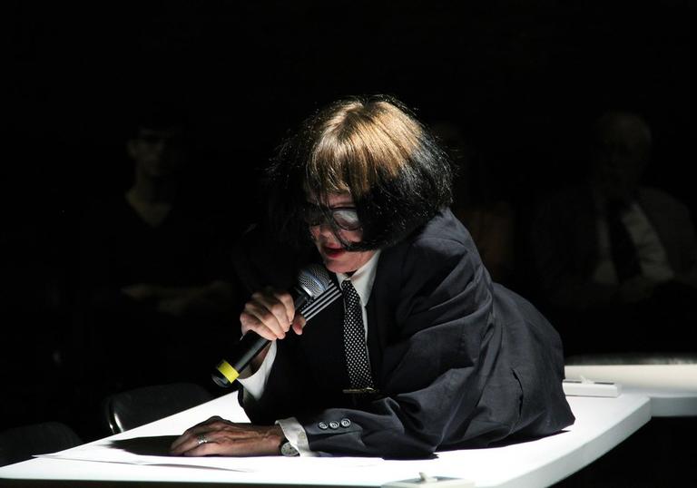 Photo of a woman in glasses using a mic. She is lying on a table. 