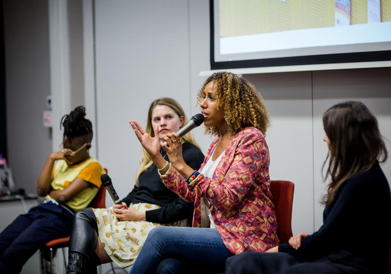 Four people on a panel, one talking into the microphone and gesticulating with a screen behind them.