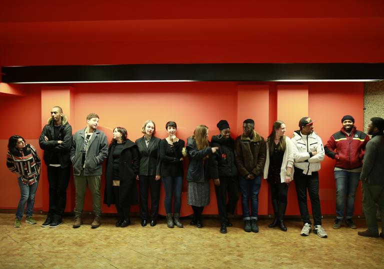 13 people standing in a line along an orange wall, talking and laughing