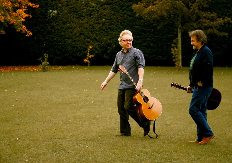 Andy Irvine and Paul Brady carrying a couple of guitars enjoying a walk on some very green grass