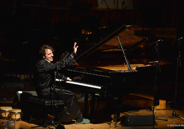 Chilly Gonzales is playing a piano one handed whilst doing a wave