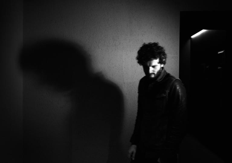 A black and white photo of Apparat in front of a wall, his shadow is cast on the wall behind him