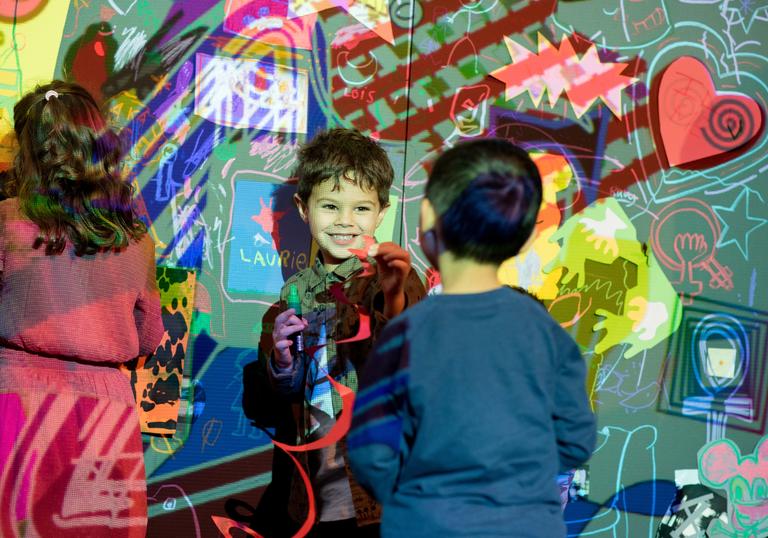 a young boy plays with his friend up against a wall projected with the work of Jean Michel Basquiat. They are both smiling and having a really nice time