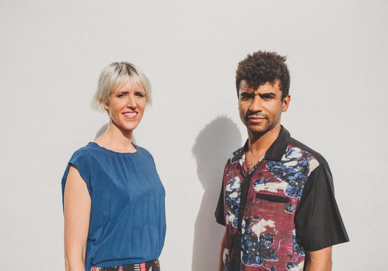 Kate Simko and Jamie Jones smile awkwardly in front of an off-white wall