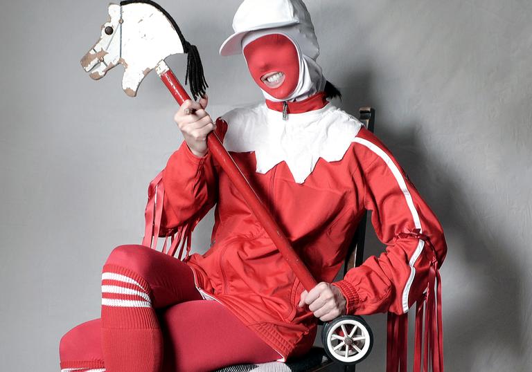 Gazelle Twin wearing a jester costume with a hobby horse, seated on a chair