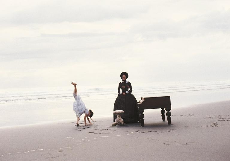 A scene from The Piano. A child does a handstand and a woman sits at a piano on a beach