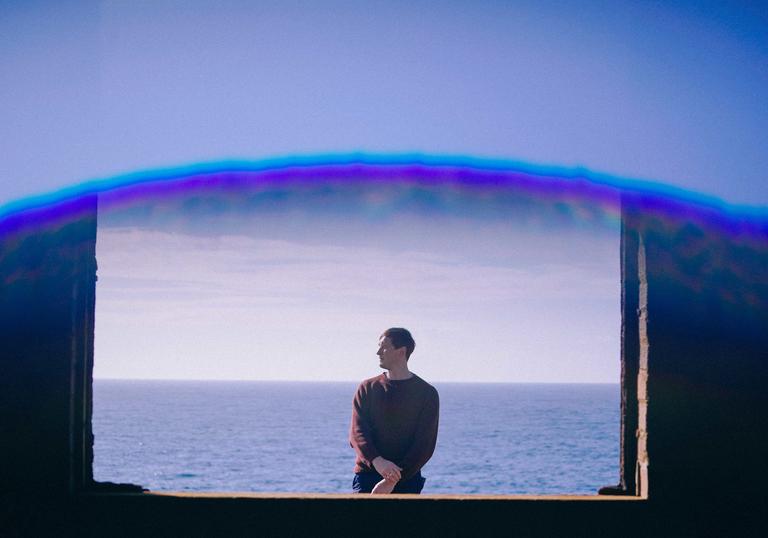 Erland Cooper standing in front of the sea, as seen through a window