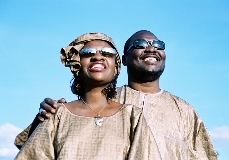 Amadou & Mariam in front of a blue sky. They are both wearing gold clothing and sunglasses.