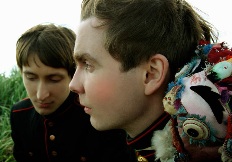 Jonsí & Alex, in military style jackets and feathered adornments, sit in long grass