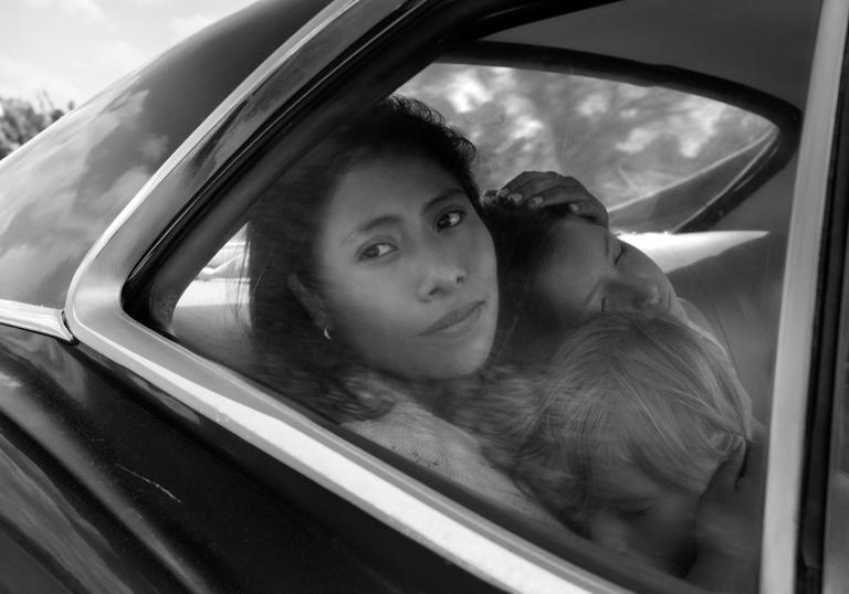 A woman hold a child in the back seat of a car and looks longingly out of the window. The image is a still from the film Roma
