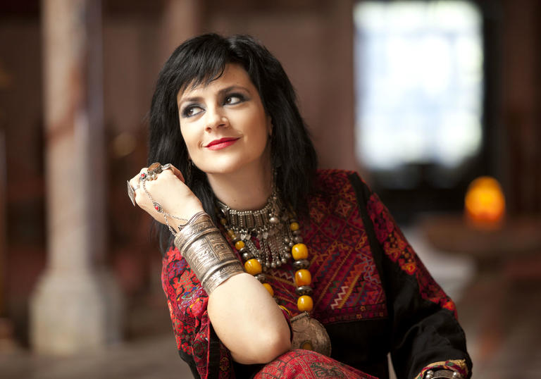 Rim Banna wearing a red dress and large jewellery. She is leaning on her elbow and looking away from the camera.