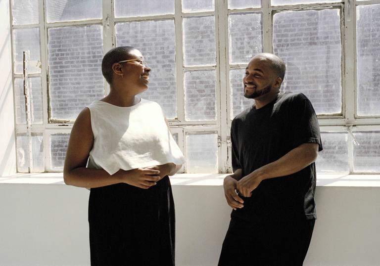 Cecile McLorin Salvant and Sullivan Fortner leaning on a ledge in front of windows with sun streaming in