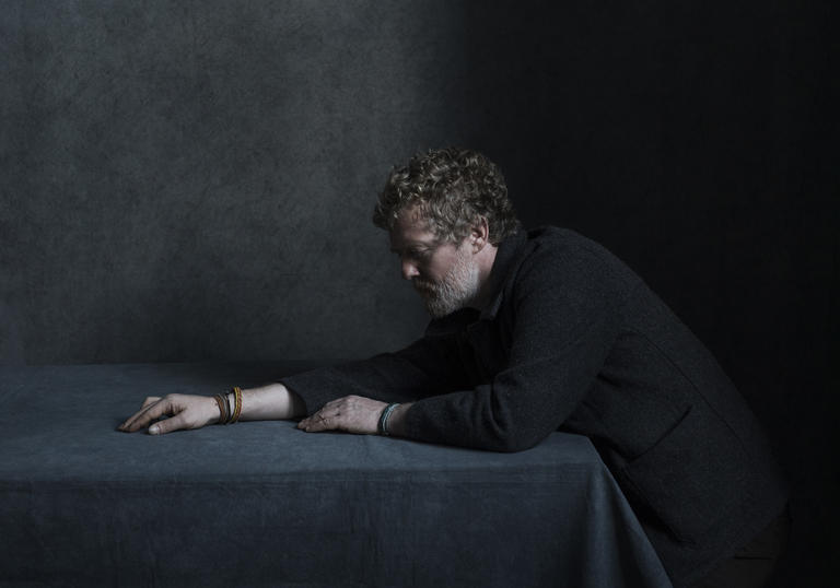 Glen Hansard stretching his right arm across a table. He is wearing leather bracelets.