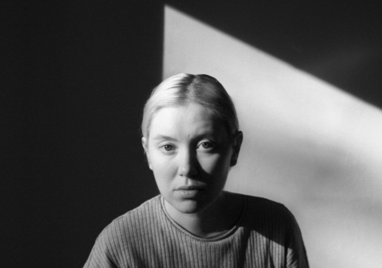 A black and white photo of Ellen Arkbro's head and shoulders. She is looking at the camera with a beam of light behind her.