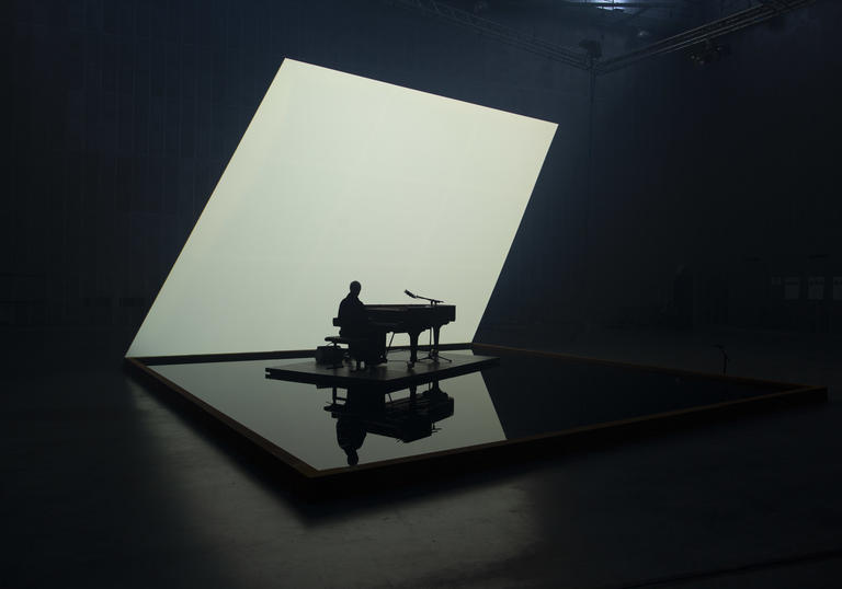 Nik Bartsch playing solo piano on a pool of water, with a lit white backdrop