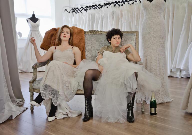 Holliday Granger and Alia Shawkat sitting in a wedding dress shop wearing wedding dresses pulled up to their knees