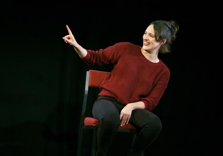 Phoebe Waller-Bridge sitting on a chair in a dark space with her arm and finger pointing up