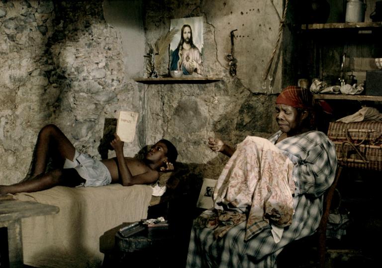 A young boy reads on a bed while his grandmother sews, in a bare-walled room in Martinique
